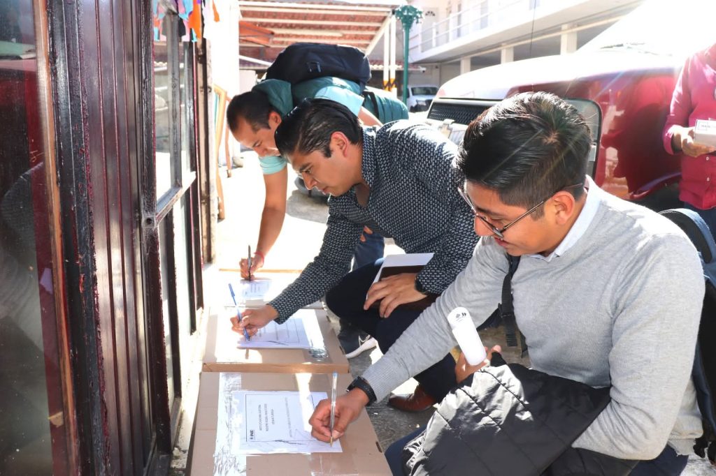 1. Personas firmando en una mesa con cajas de documentación afuera de un establecimiento.