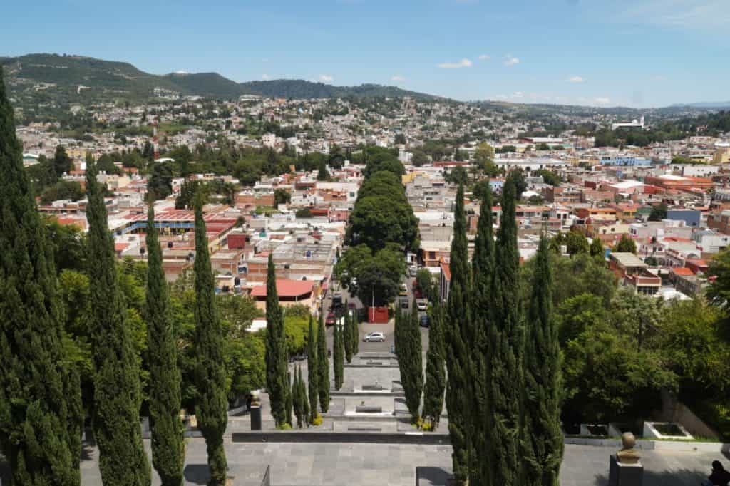 1. Vista panorámica de la ciudad de Tlaxcala con calle arbolada en primer plano y barrios en las colinas al fondo, cielo despejado y ambiente urbano.