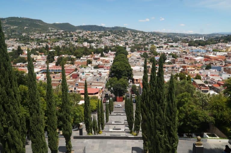 1. Vista panorámica de la ciudad de Tlaxcala con calle arbolada en primer plano y barrios en las colinas al fondo, cielo despejado y ambiente urbano.