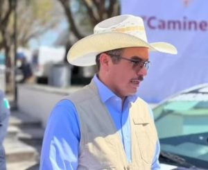 1. Hombre con sombrero y gafas, usando camisa azul y chaleco, en un entorno urbano con árboles y vehículos al fondo.