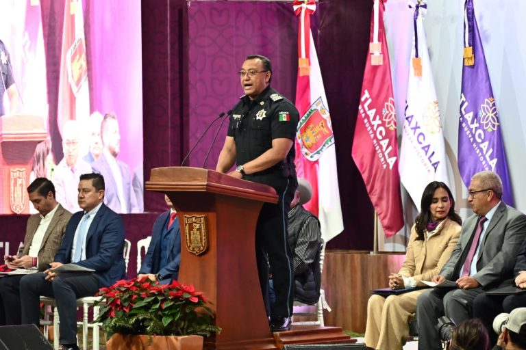 1. Oficial de policía hablando en un podio durante una ceremonia en Tlaxcala, con banderas y personas sentadas a su lado.