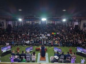 1. Gran evento con una gran multitud en un salón, con una oradora en un podio, rodeada de mesas con autoridades y pantallas que muestran la transmisión del evento.