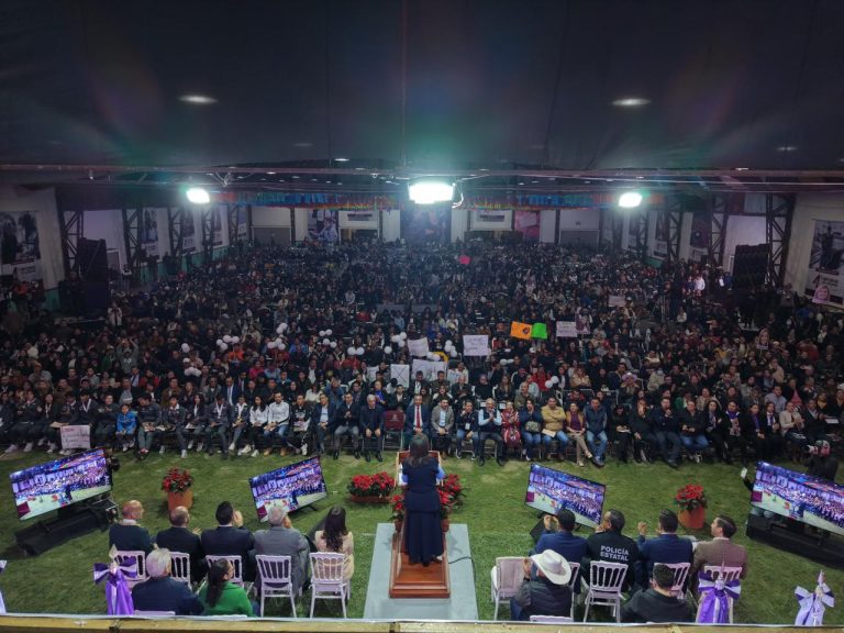 1. Gran evento con una gran multitud en un salón, con una oradora en un podio, rodeada de mesas con autoridades y pantallas que muestran la transmisión del evento.