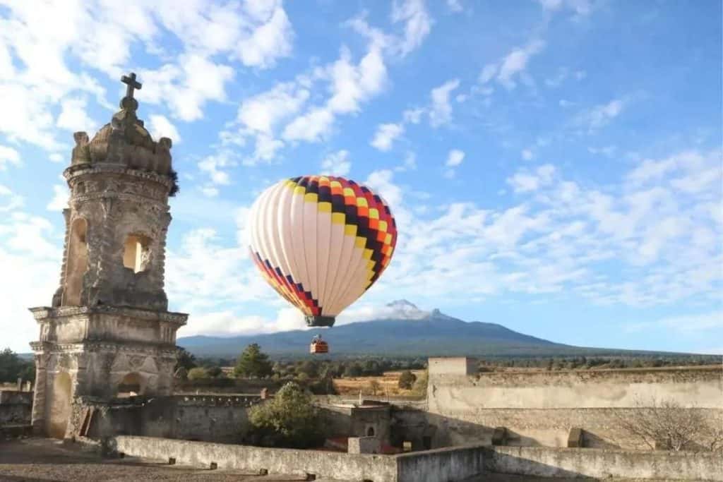 1. Globo aerostático sobre un paisaje con torre antigua y montañas al fondo, cielo con nubes dispersas en un día despejado en Tlaxcala, México.