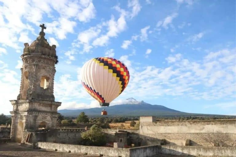 1. Globo aerostático sobre un paisaje con torre antigua y montañas al fondo, cielo con nubes dispersas en un día despejado en Tlaxcala, México.