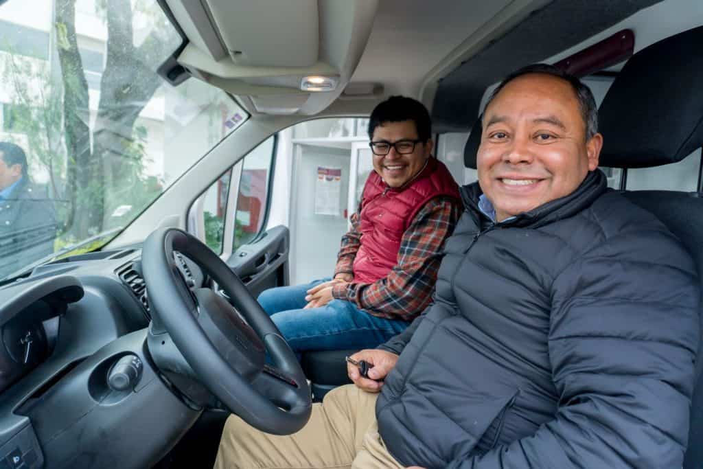 1. Dos hombres sonrientes en la cabina de una van, uno con gafas y vestimenta casual, con ambiente urbano y natural visible a través del parabrisas.