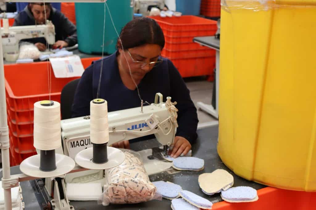 Personal trabajando con máquina de coser en un almacén o fábrica, rodeada de cajas y materiales de trabajo, en un entorno industrial.