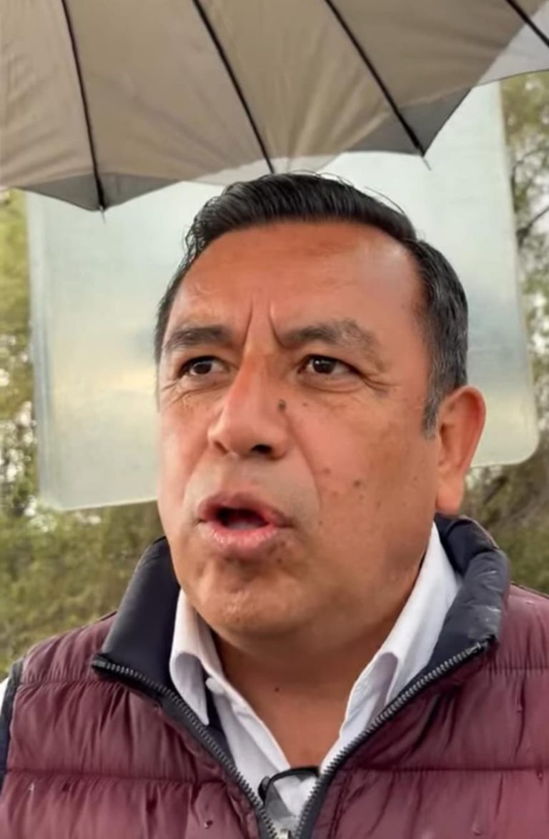1. Hombre con paraguas en exteriores, expresión seria, en un ambiente natural con árboles, usando chaqueta y camisa blanca.