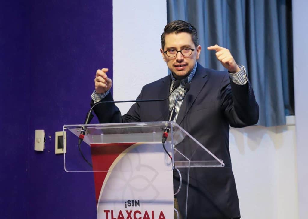 1. Hombre en traje y gafas, dando un discurso en un podio con fondo de cortinas azules y paredes blancas, en un evento formal en Tlaxcala, México.
