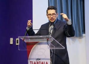 1. Hombre en traje y gafas, dando un discurso en un podio con fondo de cortinas azules y paredes blancas, en un evento formal en Tlaxcala, México.