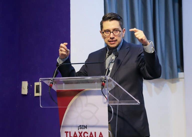 1. Hombre en traje y gafas, dando un discurso en un podio con fondo de cortinas azules y paredes blancas, en un evento formal en Tlaxcala, México.