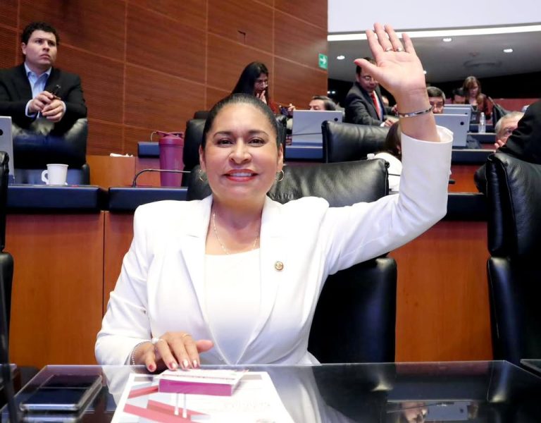 1. Mujer en una sesión parlamentaria, levantando la mano y sonriendo, vestida con traje blanco, en un entorno formal y profesional.