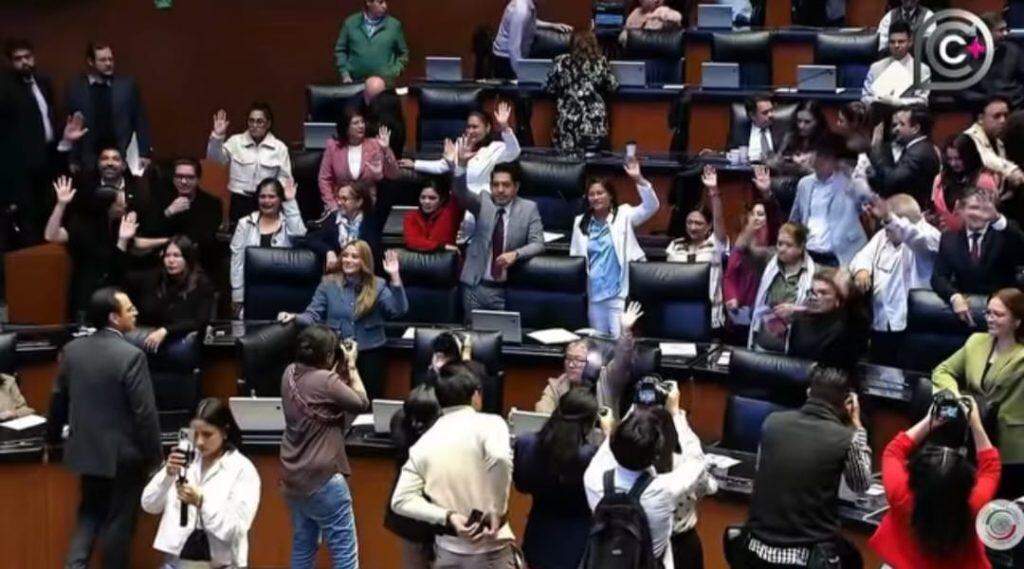 Diputados en la LXIV Legislatura de México en sesión plenaria, algunos levantando la mano, otros tomando fotos y conversando en el recinto parlamentario.