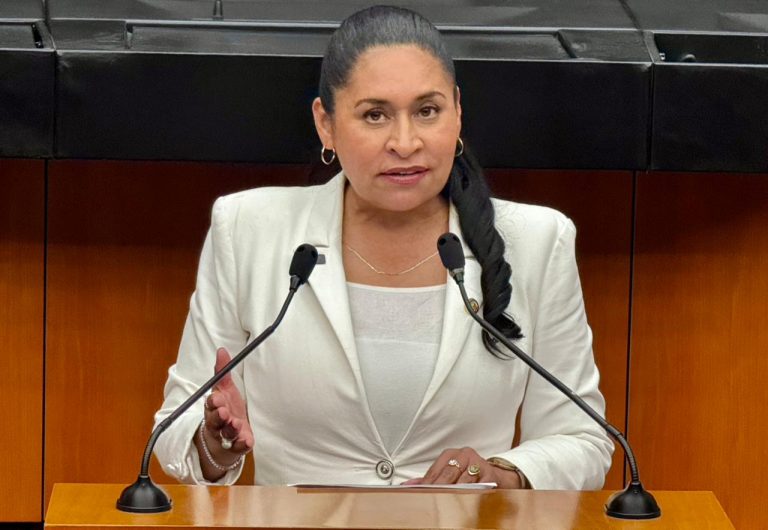 1. Mujer política en una sala de prensa, hablando en micrófonos, vestida con saco blanco, expresando autoridad y liderazgo.