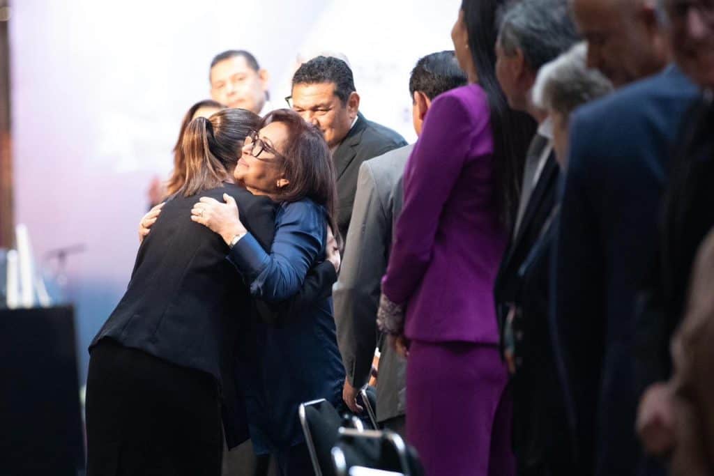 1. Mujer abrazando a otra mujer en un evento formal, rodeada de personas en línea, con expresión de afecto y apoyo.