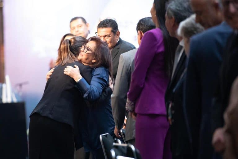 1. Mujer abrazando a otra mujer en un evento formal, rodeada de personas en línea, con expresión de afecto y apoyo.