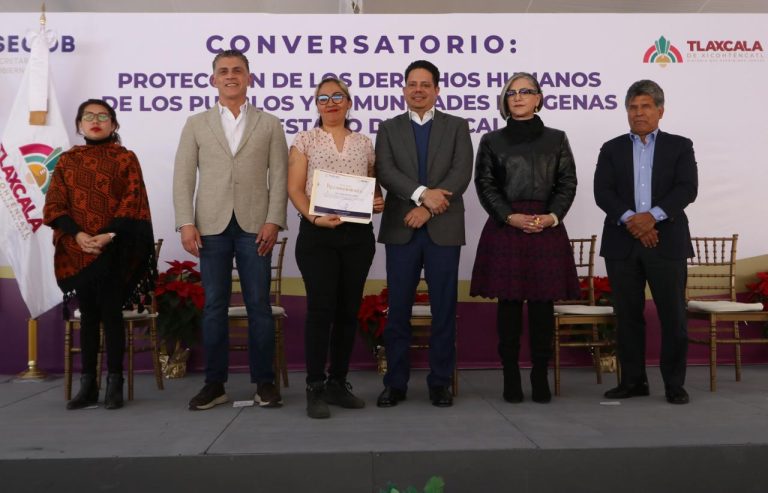1. Mujeres y hombres en ceremonia, algunos sosteniendo un reconocimiento, frente a un cartel sobre derechos humanos y protección a comunidades indígenas en Tlaxcala.