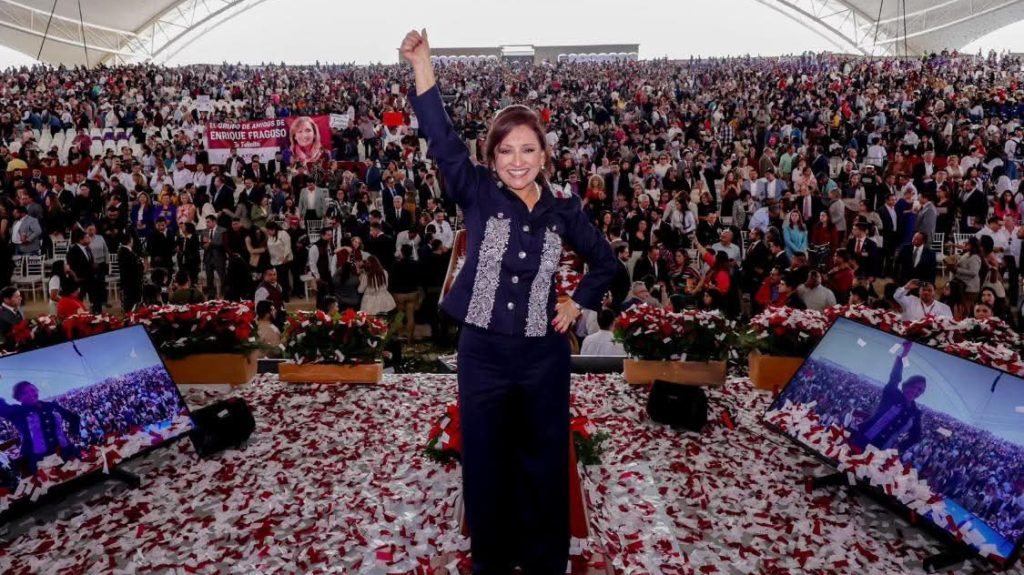 1. Ilustración de una mujer celebrando en un gran evento con multitud de personas en un estadio cubierto, rodeada de pantallas y confeti en el suelo, símbolo de éxito y liderazgo.