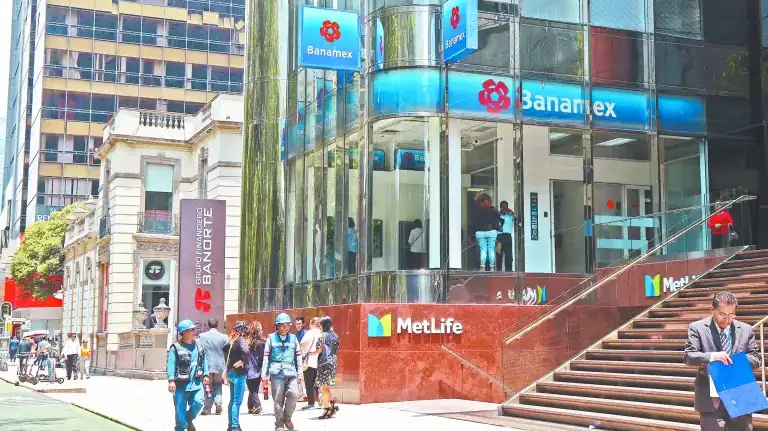 Personas caminando frente a la sucursal de Banamex en una calle del centro de la ciudad, con edificios modernos y tradicionales en el entorno urbano.