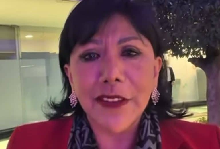 Blanca Juárez, una mujer de cabello corto y oscuro, usa un blazer rojo y accesorios de playa en pendientes y un pañuelo con patrón, en una escena urbana con árboles y un edificio de fondo.
