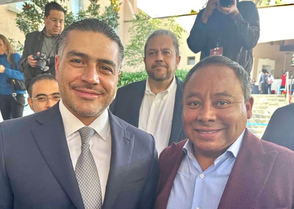 1. Dos hombres en traje toman una selfie en un evento oficial, rodeados de otras personas y fotógrafos en un ambiente al aire libre.