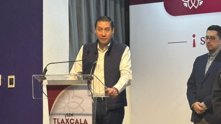 1. Hombre con chaqueta negra y camisa blanca dando un discurso en un podio con fondo blanco y logo de prensa en Tlaxcala, México.