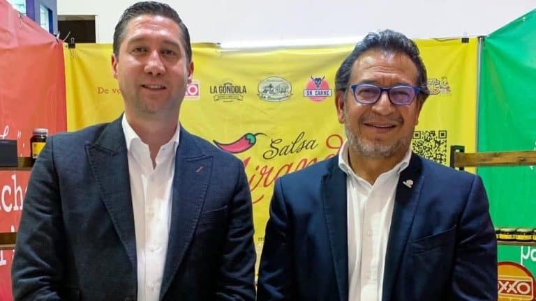 1. Dos hombres con trajes oscuros y gafas, sonriendo en un evento, con fondo de banner amarillo y verde que promociona salsa y otros productos locales de Tlaxcala, México.
