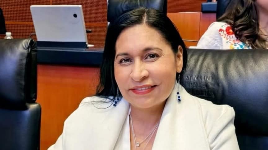 Profesional woman smiling at a formal event, seated on a black leather chair, with a wooden background and a laptop behind her.