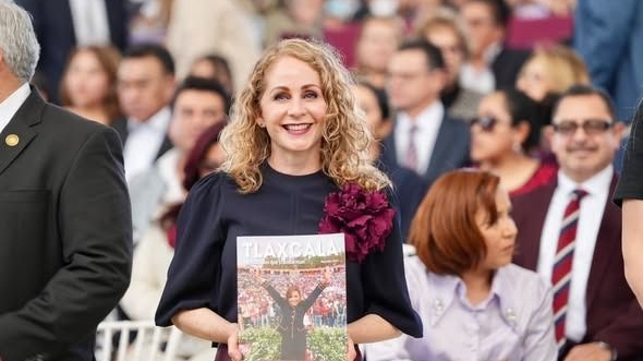 Una mujer sonriendo en un evento público, sosteniendo una revista de Tlaxcala con fondo de personas vestidas formalmente.