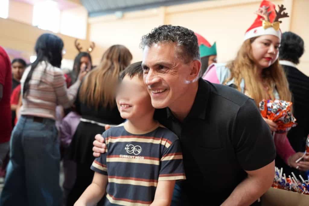 1. Un hombre sonriente abrazando a un niño en una celebración navideña con personas vestidas con gorros y accesorios festivos en un ambiente lleno de alegría.
