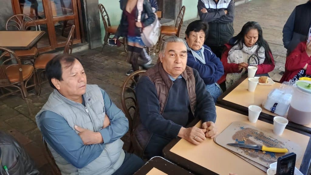 Hombres y mujeres adultos sentados en una mesa en un restaurante informal, algunos con expresiones serias, tomando comida y bebida, en un entorno acogedor de Tlaxcala, México.