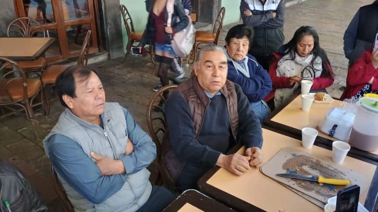 Hombres y mujeres adultos sentados en una mesa en un restaurante informal, algunos con expresiones serias, tomando comida y bebida, en un entorno acogedor de Tlaxcala, México.
