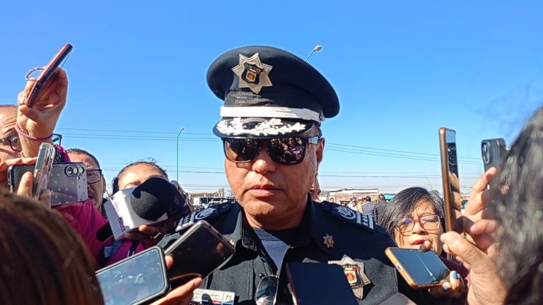 1. Oficial de policía con gorra y lentes oscuros rodeado de periodistas y medios de comunicación en exteriores bajo cielo azul.