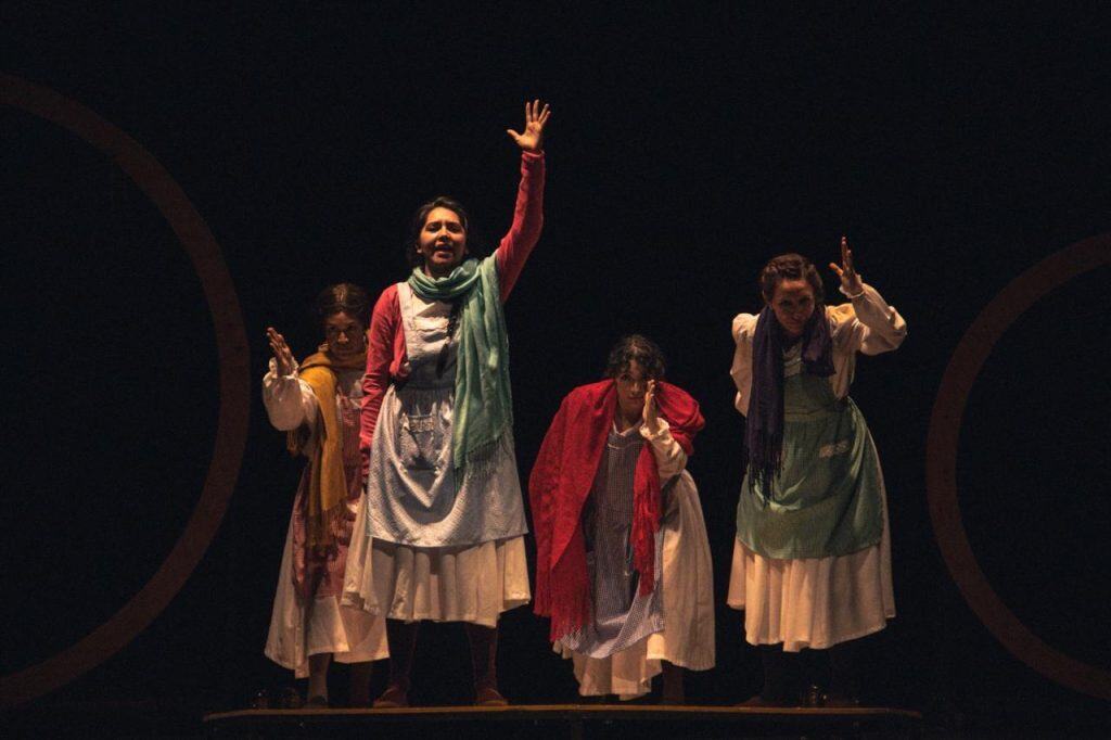 Una escena de una obra teatral con cuatro actrices en escenario oscuro, vestidas con prendas tradicionales y expresando emociones dramáticas durante una presentación cultural en Tlaxcala.