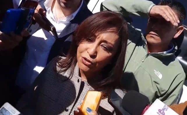 1. Mujer con cabellera castaña, rodeada de periodistas y micrófonos, en una apariencia de entrevista o declaración pública.