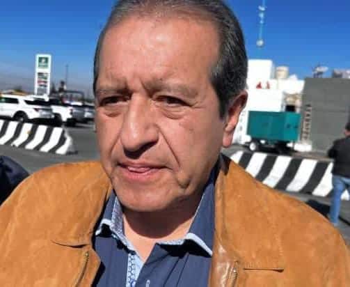 1. Hombre de rostro serio con cabello gris, lleva chaqueta marrón y camisa con cuello abierto, en un entorno urbano con cielo azul y estructuras en el fondo.