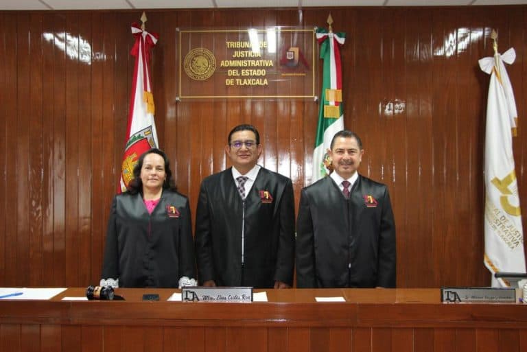 Jueces en traje en una sala de audiencia con banderas de Tlaxcala y un cartel que dice "Tribunal de Justicia Administrativa del Estado de Tlaxcala".