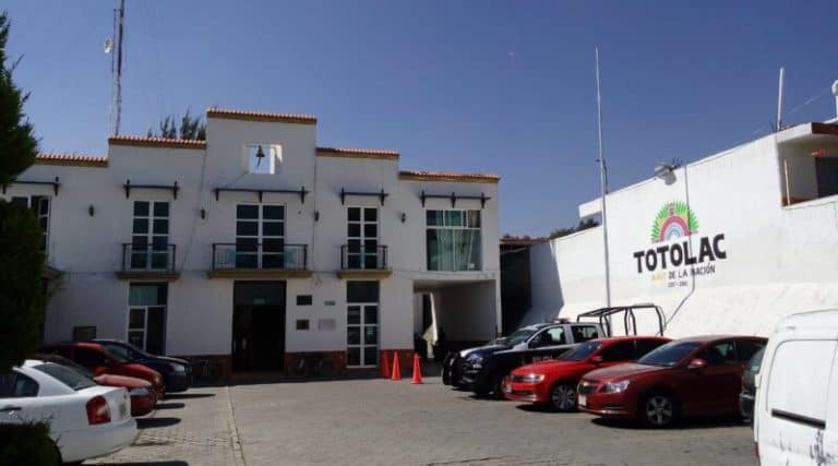 Atractiva fachada del edificio de Totolac, Tlaxcala, con varios autos estacionados en el frente y un mural que destaca en la pared. El cielo despejado y el entorno reflejan un día soleado en el municipio.