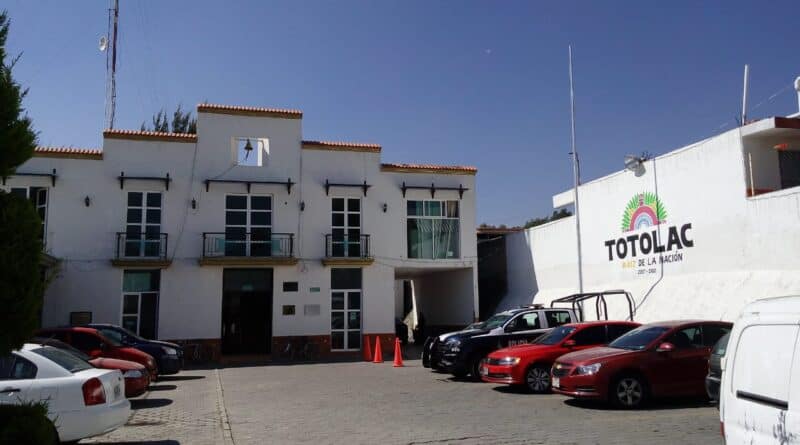Atractiva fachada del edificio de Totolac, Tlaxcala, con varios autos estacionados en el frente y un mural que destaca en la pared. El cielo despejado y el entorno reflejan un día soleado en el municipio.