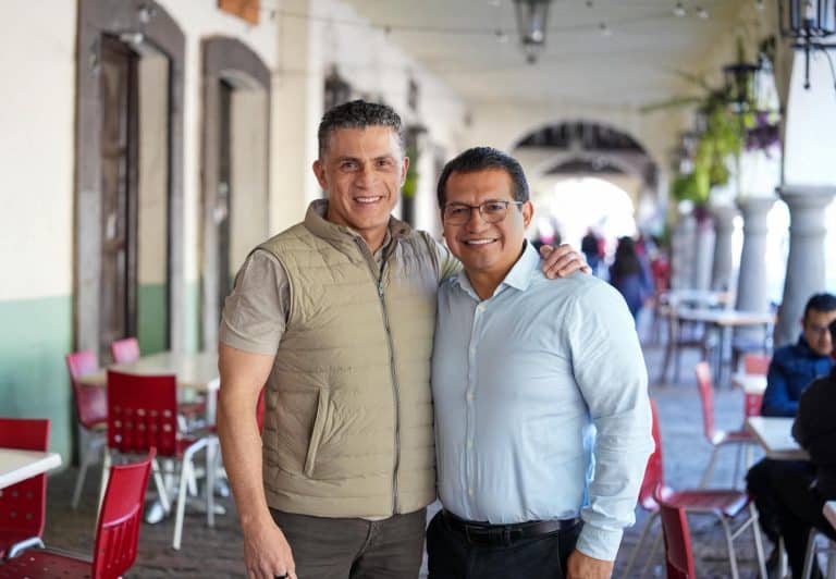 1. Dos hombres sonriendo, uno con chaleco beige y otro con camisa blanca, en un espacio abierto con arcadas y mesas rojas.