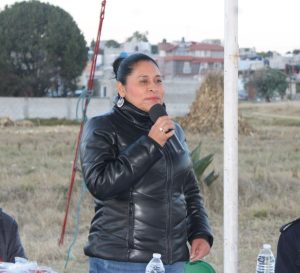 Mujer con cabello recogido, vestida con chaqueta de cuero negra, hablando con micrófono en mano en evento al aire libre en Tlaxcala.