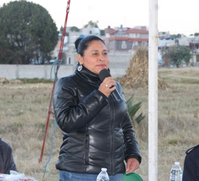Mujer con cabello recogido, vestida con chaqueta de cuero negra, hablando con micrófono en mano en evento al aire libre en Tlaxcala.
