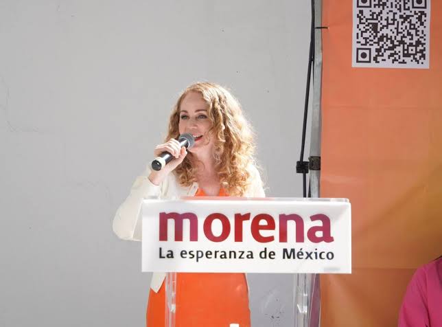 Una mujer con cabello rizado y rubio hablando en micrófono en un evento político, con un cartel de Morena que dice "La esperanza de México".