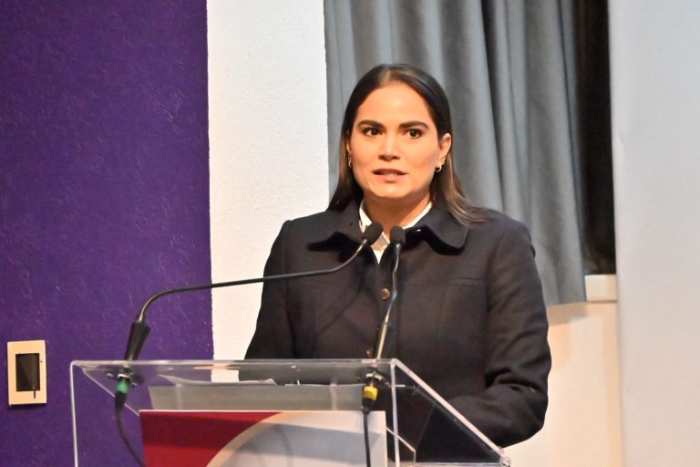 Innovadora mujer en un discurso público en un evento formal, con fondo de cortinas grises, vestida con chaqueta negra y camisa blanca, transmitiendo autoridad y confianza.
