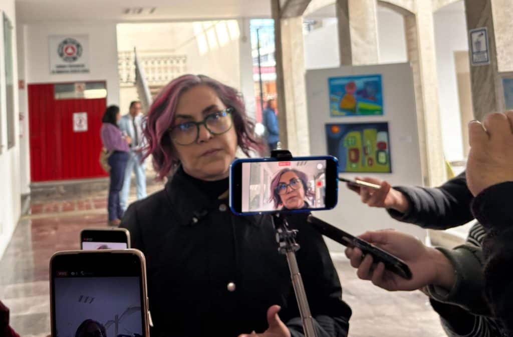 1. Mujer con cabello rosado y gafas siendo entrevistada con teléfonos y micrófono en un espacio interior, en medio de una cobertura periodística en Tlaxcala, México.
