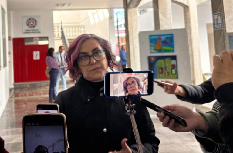 1. Mujer con cabello rosado y gafas siendo entrevistada con teléfonos y micrófono en un espacio interior, en medio de una cobertura periodística en Tlaxcala, México.