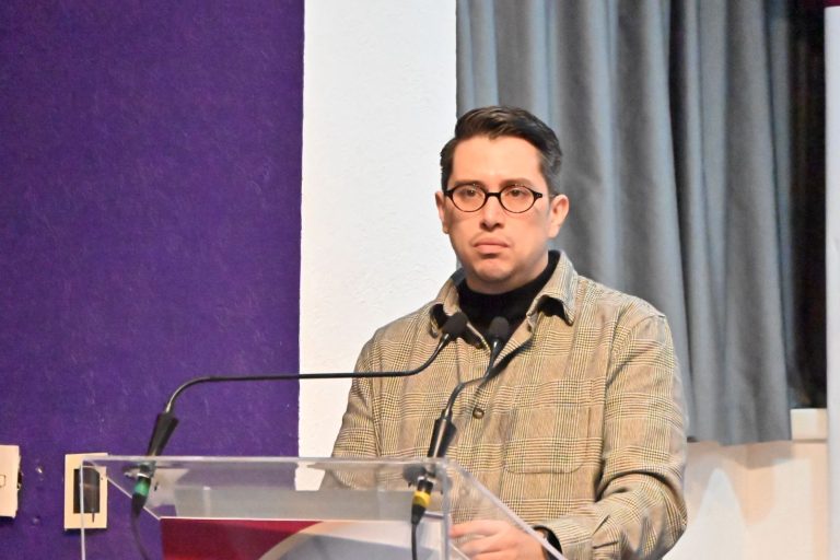 La segunda persona, con gafas y traje a cuadros, hablando en un podio durante un evento formal en un espacio cerrado con cortinas grises.