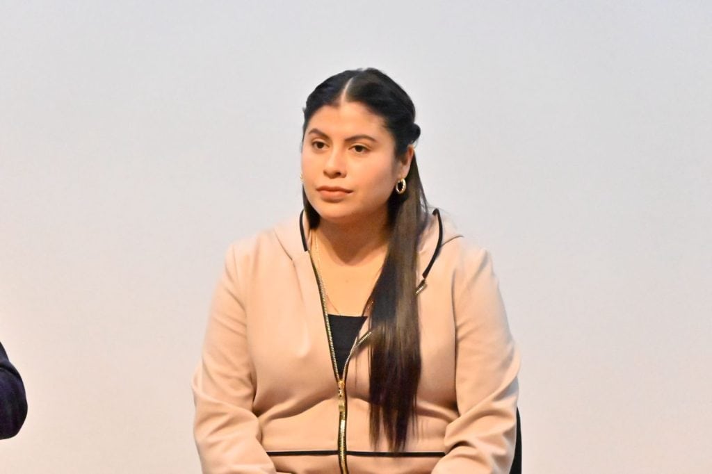 Parece que la imagen muestra a una mujer en una reunión o conferencia, con expresión seria, vestida con un blazer beige y cabello liso recogido.
