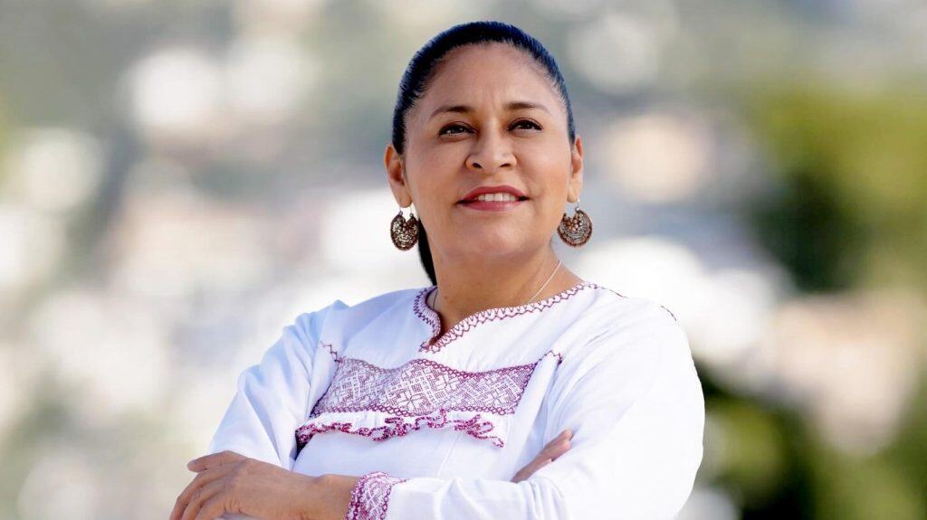 1. Mujer tlaxcalteca con peinado recogido, vestida con blusa blanca y detalles en fucsia, posa con confianza en un entorno natural al aire libre.