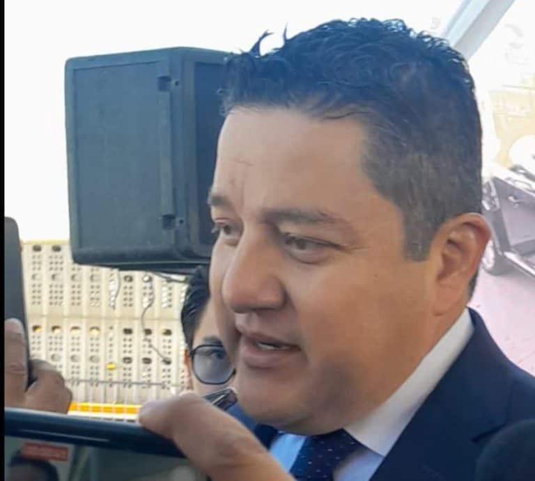 1. Hombre con traje y corbata hablando frente a micrófono y cámara, en un evento, con fondo de equipo de sonido y estructura metálica.